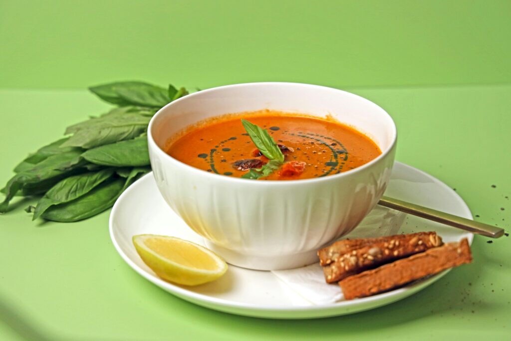 pexels-photo-14774695-14774695 A vibrant tomato soup garnished with basil, accompanied by lemon and breadsticks.