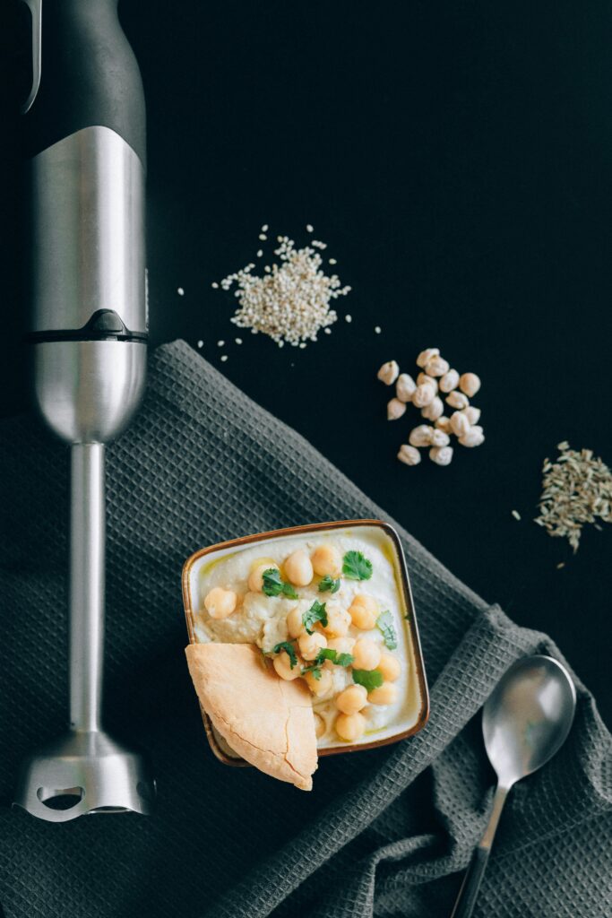 pexels-photo-6252742-6252742 Flatlay of creamy hummus with chickpeas and pita, surrounded by blender and herbs.